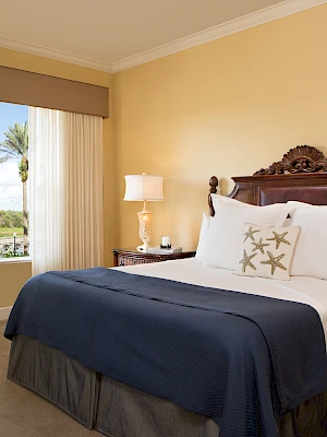 A cozy bedroom features a wooden bed with white and blue bedding, a floral chair, side tables with lamps, and a window view of palm trees.