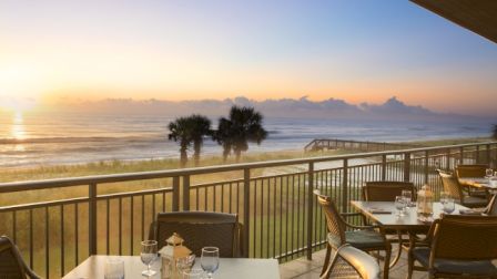 A scenic beachfront dining area with set tables, overlooking the ocean and palm trees at sunrise or sunset.