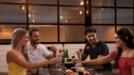 Four people enjoying sushi and drinks at a restaurant, smiling and engaging in conversation in a warmly lit setting.
