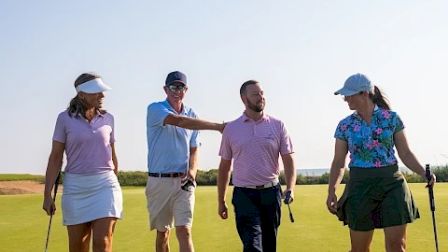 A group of four friends walking on a sunny golf course, carrying golf clubs, enjoying a day outdoors.