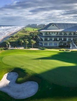A seaside golf course with a manicured green, sand traps, and a large building nearby by the ocean, under a partly cloudy sky.