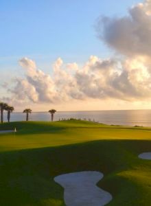 A sunny coastal golf course with green fairways, sand bunkers, a calm sea, a few palm trees, and dramatic clouds at sunset, ending gracefully.