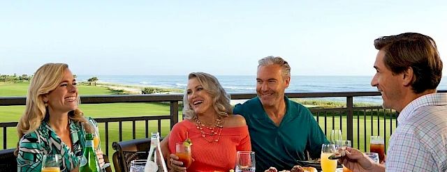 A group of four adults enjoys a sunny outdoor meal on a balcony with ocean view, sharing drinks and plates of food.