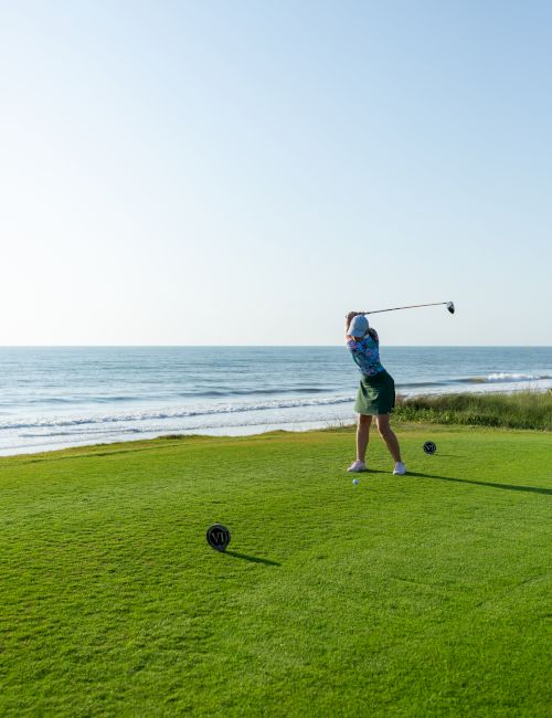 A person playing golf swings on a grassy course by the ocean, under a clear blue sky.