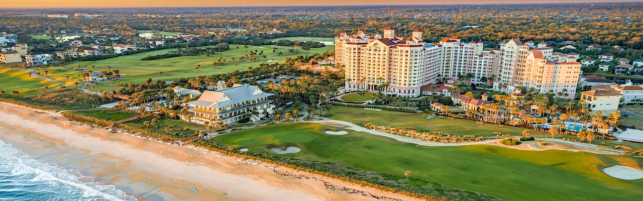 Aerial view of a coastal resort with large buildings, a golf course, and a sandy beach along the ocean.