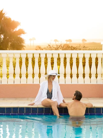 Two people lounging by a pool at sunset, a woman in a hat and cover-up talking to a man in the water, with a pastel terrace in the background.