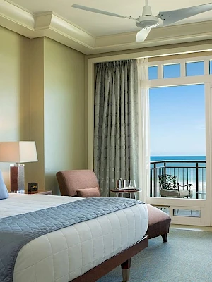 A hotel room with a bed, lamps, chairs, and a balcony offering an ocean view can be seen in the image.