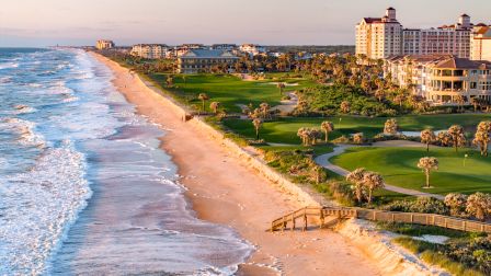 The image shows a coastal scene with waves, sandy beach, and buildings set against a lush green landscape under a clear sky.