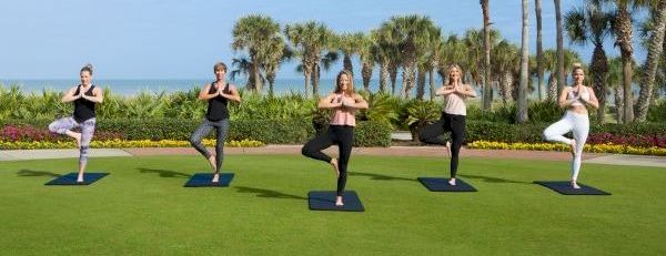 Five people do yoga on mats outdoors, palm trees and blue sky in a tropical setting.