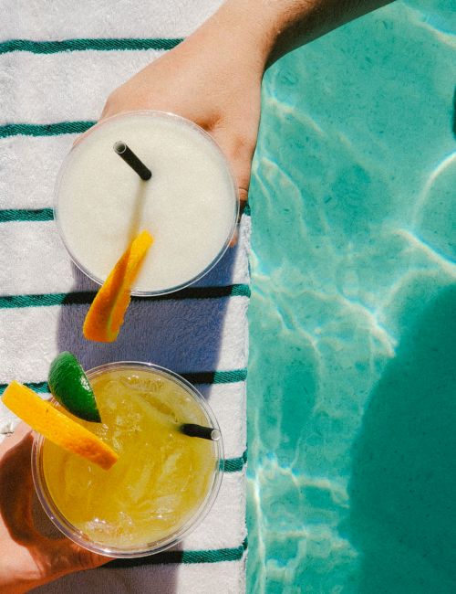 Two drinks with straws and citrus garnishes sit on a striped towel beside a pool.
