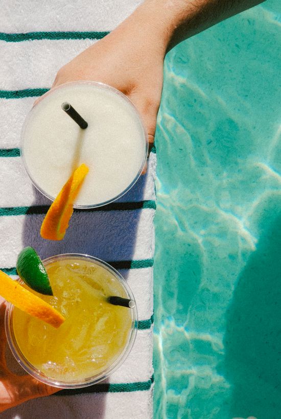 Two drinks with straws and citrus garnishes sit on a striped towel beside a pool.