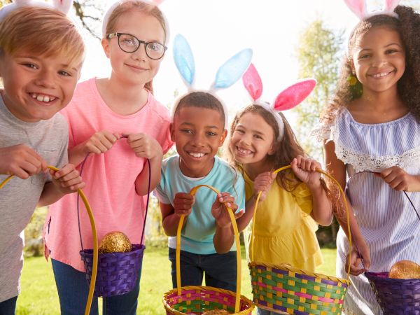 Five children with bunny ears hold colorful baskets with golden eggs, smiling outdoors in the sunlight.