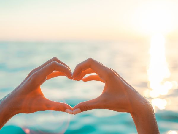 Two hands form a heart shape against a sunset over the water, creating a romantic seaside vibe.