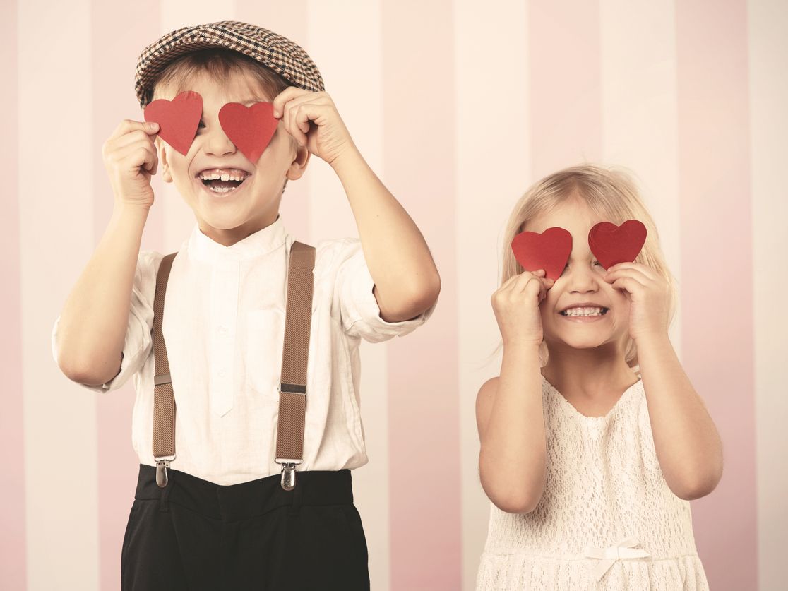 Two kids smile and hold red heart shapes over their eyes, posing playfully in front of pink striped backdrop.