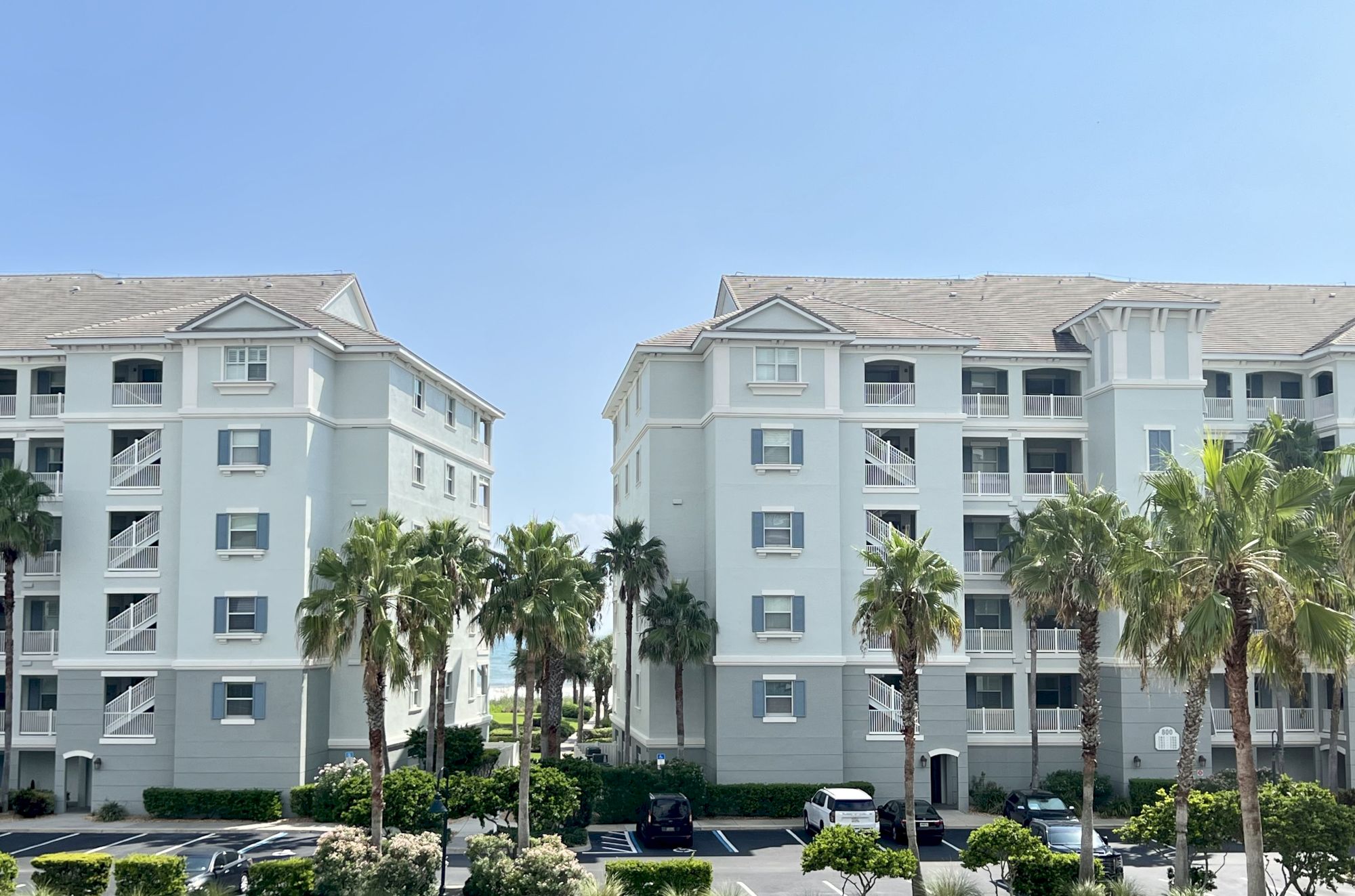 The image shows two light-colored apartment buildings with palm trees in front, a parking area, and a clear blue sky.
