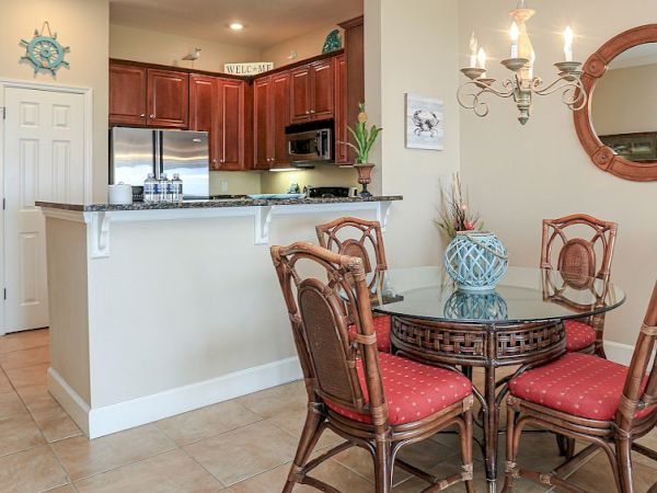 This image shows a cozy kitchen and dining area with wooden cabinets, a round table with red-cushioned chairs, and a decorative chandelier.