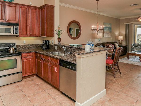 The image shows a kitchen with wooden cabinets, stainless steel appliances, and granite countertops, leading to a dining and living area.