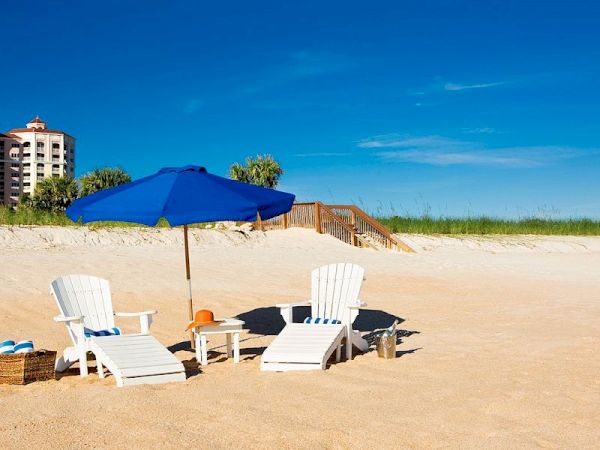 Two white beach chairs, a blue umbrella, and a basket are on a sandy beach with clear skies and some buildings in the background.