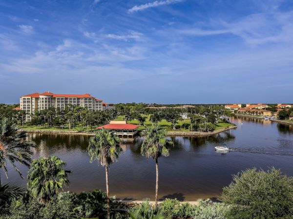 Scenic view of a resort with buildings, palm trees, and a boat on a calm body of water under a clear blue sky.