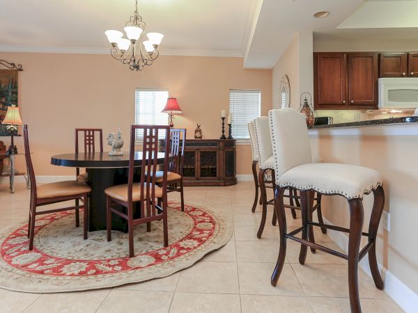 A cozy dining room with a round table, four chairs, bar stools, and warm lighting, featuring a decorative rug and wooden furniture.