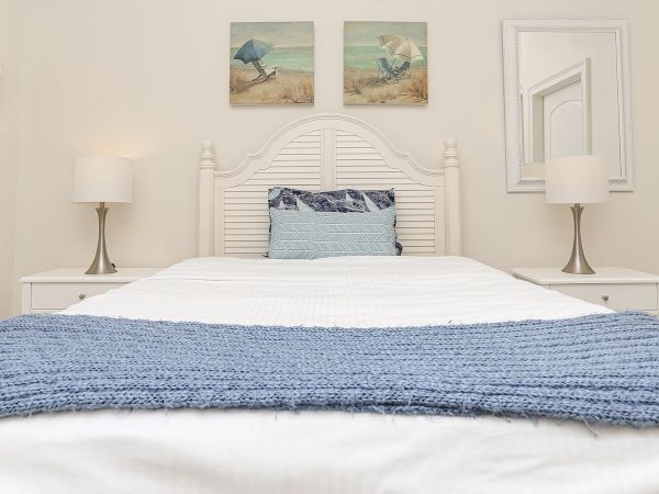 A neatly made bed with blue and white bedding, flanked by lamps on side tables. Beach-themed art and a mirror adorn the walls.