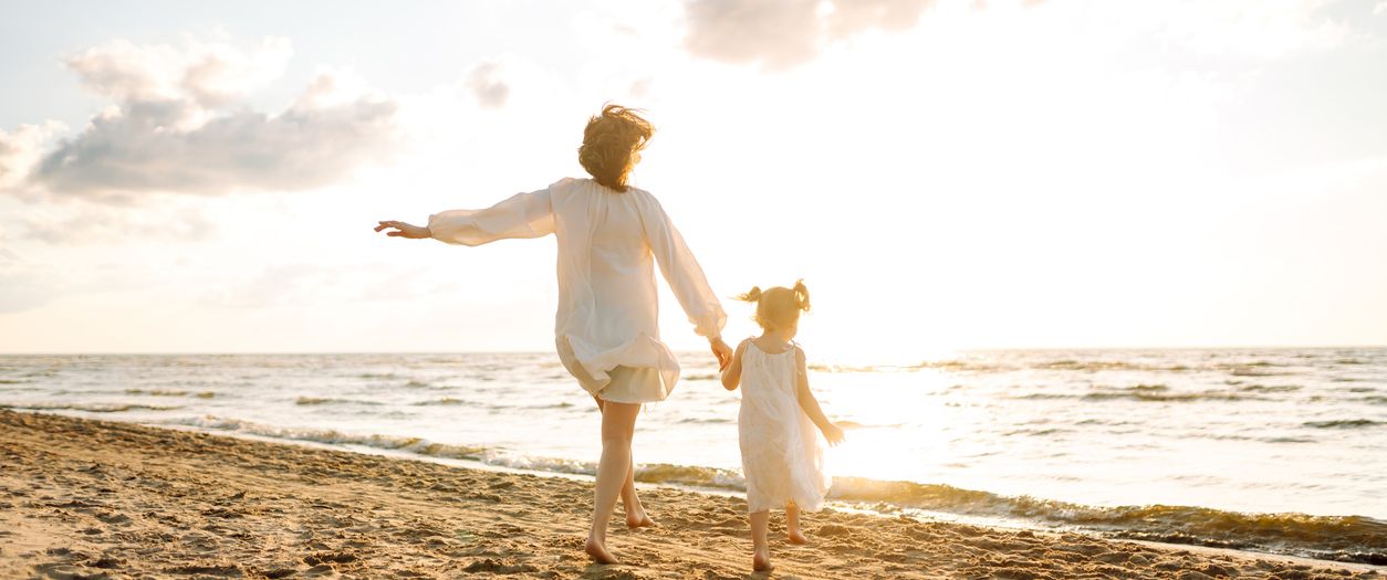 Two people, likely a parent and child, walk hand-in-hand along a sunny beach at the shore, enjoying the open horizon and waves.