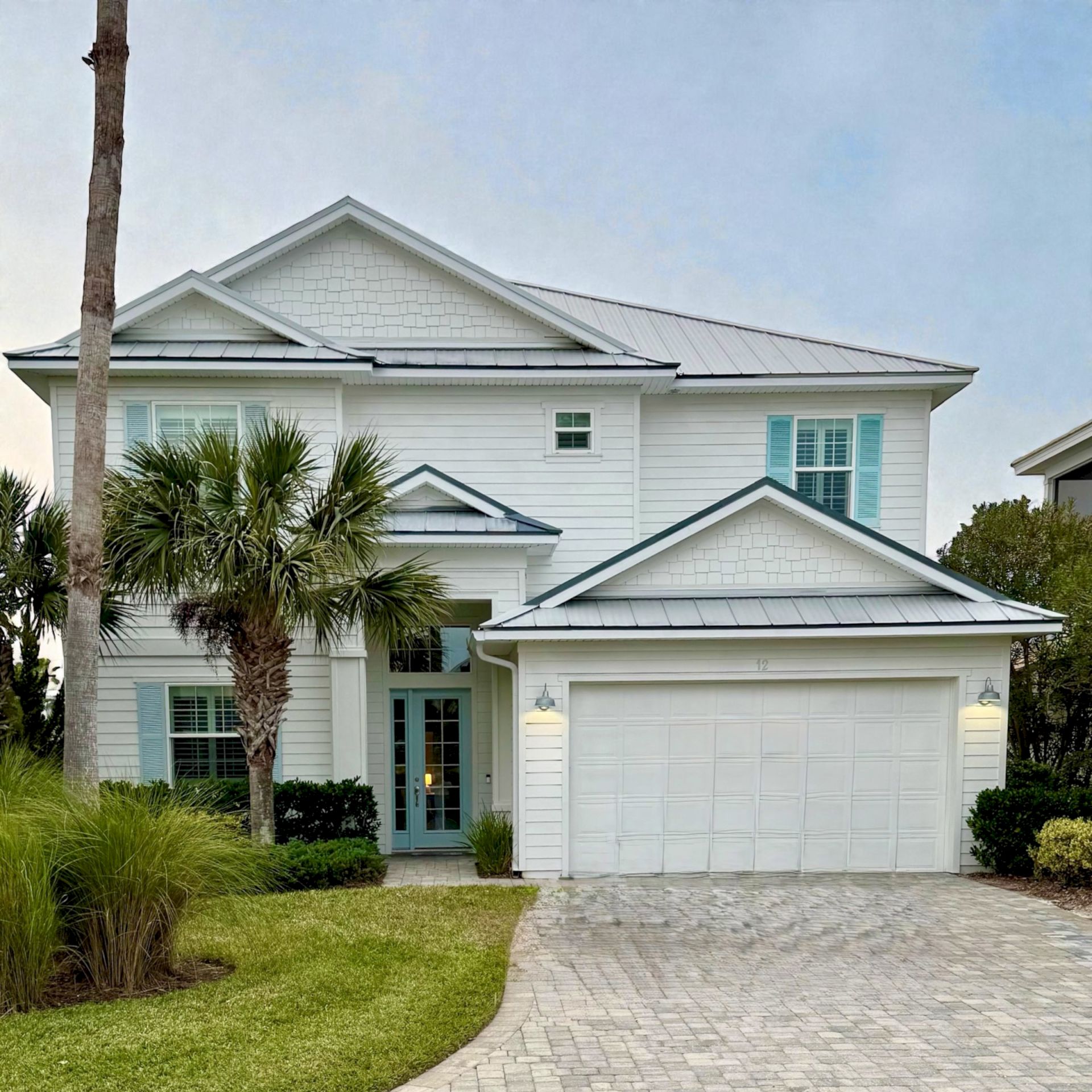 A white house with a metal roof, bay windows, and a brick walkway, framed by palm trees under a cloudy sky.
