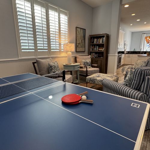 A cozy game room with a ping-pong table, snacks, and a small kitchen area in the background, plus a couch and TV.