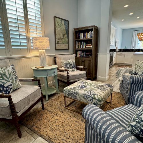 A cozy living room with beige and blue tones, a striped sofa, armchairs, a wooden coffee table, bookshelf, and warm ambient lighting.