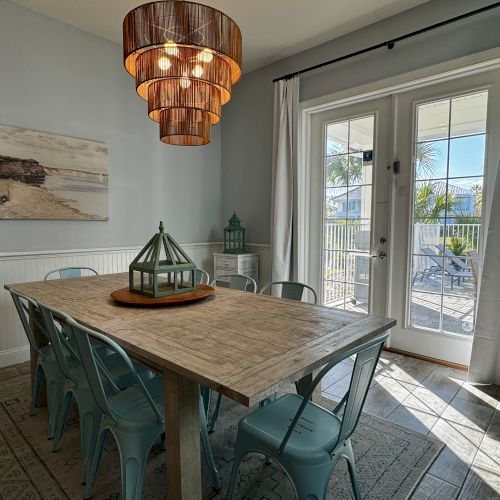 A sunlit dining room with a wooden table, teal metal chairs, a hanging pendant light, a coastal art piece, and glass doors leading to an outdoor area.