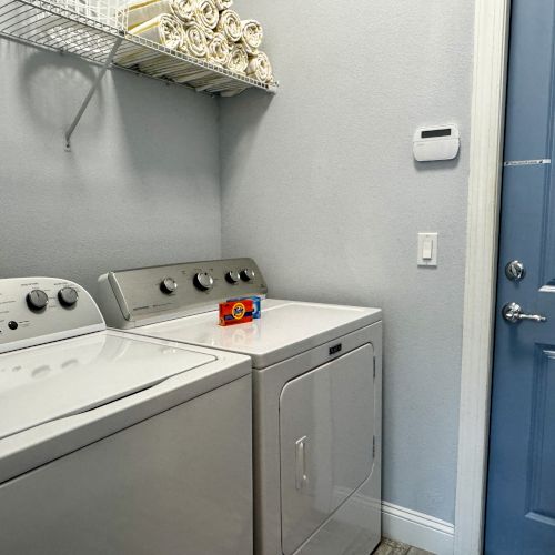 A compact laundry room with a top-loading washer and a front-loading dryer, a metal shelf above, and a blue door to the right.
