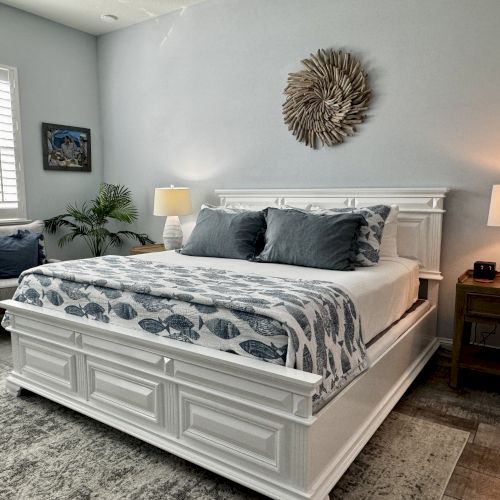 A bright, airy bedroom with a white paneled bed, gray and blue bedding, matching nightstands, lamps, a sunburst wall decor, and a cozy seating area by the window.