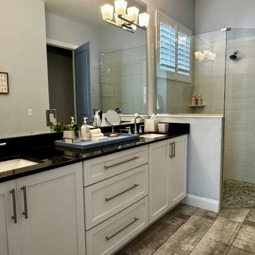 A modern bathroom with a double vanity, black countertops, white cabinets, and a glass-enclosed shower with tiled floor. End.