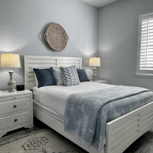 A bright, airy bedroom with a white wooden bed, matching nightstands, blue-gray bedding, and two table lamps&mdash;cozy and neatly styled.