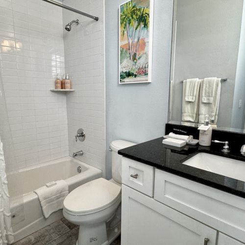 A modern bathroom with a white tub/shower, toilet, and a black countertop vanity; towels, a framed picture, and a mirror are present.