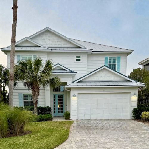 A white house with gabled roof and turquoise shutters, framed by palm trees and a bright blue sky.