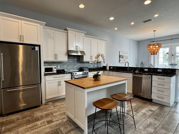 A bright, modern kitchen with white cabinets, stainless steel appliances, a central island, and two wooden stools, sunlit and organized.