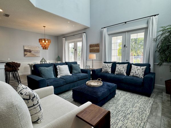 Cozy living room with a navy sectional, light gray armchairs, patterned pillows, and a large rug; bright windows and plants create a welcoming space.