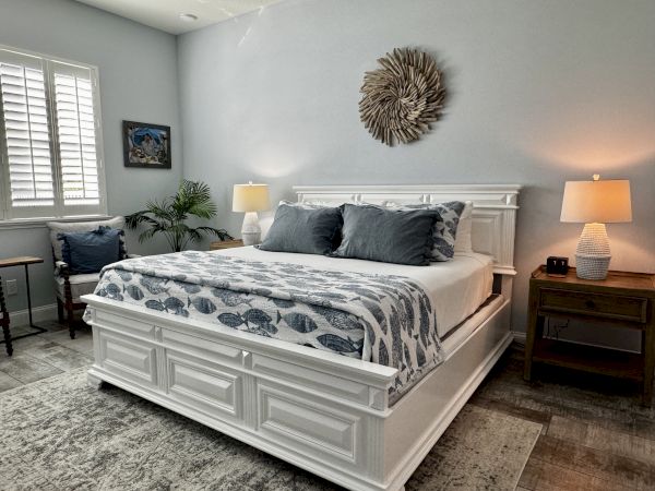 A bright, neatly made white bedroom with a large bed, blue-grey accents, two nightstands, lamps, a round wall decor, and a window with shudders.