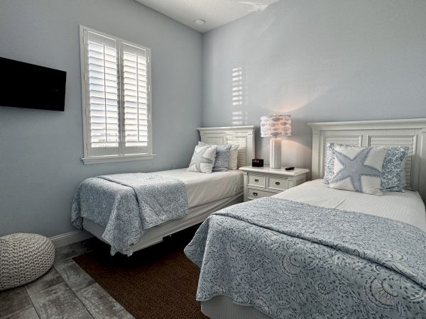 Two twin beds with light blue bedding and white headboards in a small, neatly arranged bedroom, a wall-mounted TV, a window with shutters, and a nightstand with a lamp.