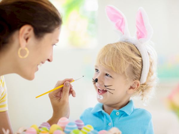 A child with bunny ears gets face paint from an adult, smiling as colorful Easter eggs decorate a table nearby.