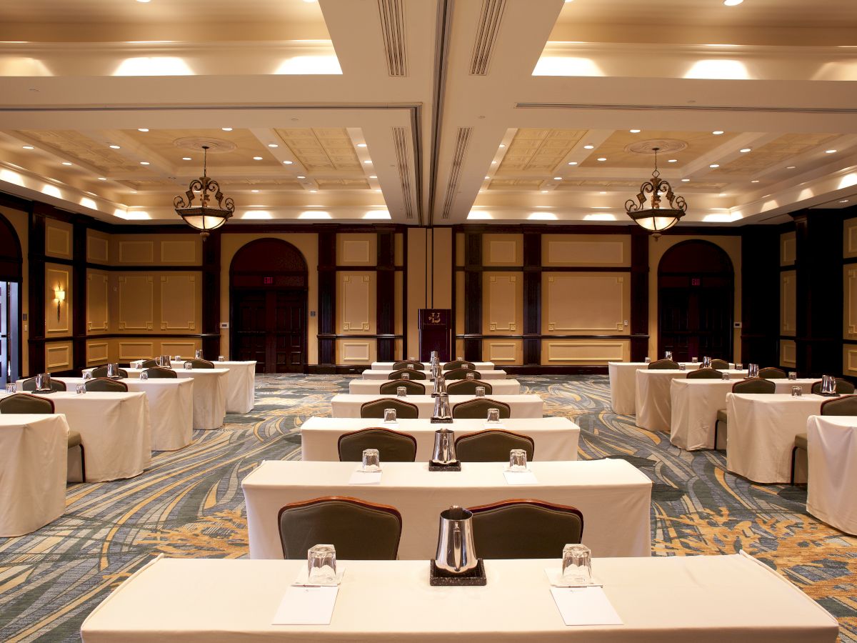 Conference room setup with long tables, white linens, and chairs arranged for a meeting or banquet in a hotel ballroom, ready for attendees.