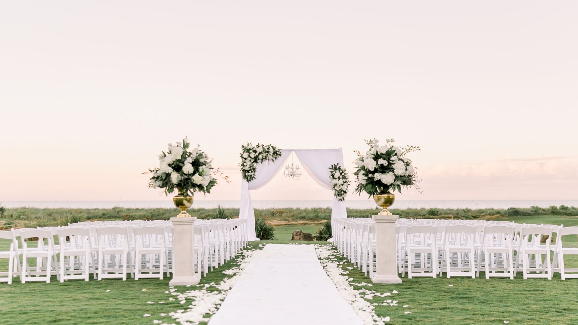A wedding setup on a grassy field with a white aisle, arch, and rows of white chairs, ready for guests to arrive. Ending the sentence.