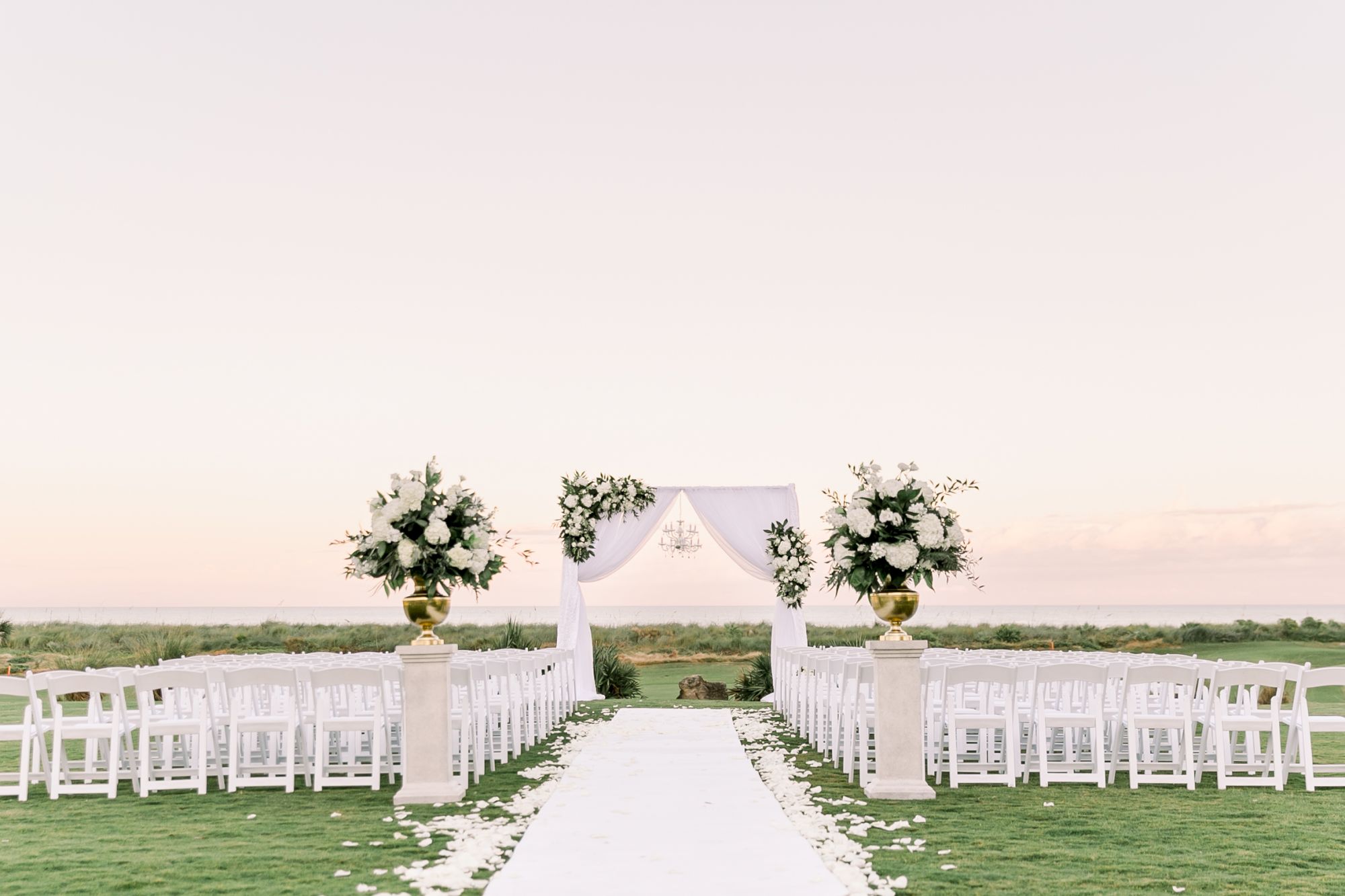 A wedding setup on a grassy field with a white aisle, arch, and rows of white chairs, ready for guests to arrive. Ending the sentence.
