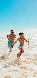 Two kids run through the gentle surf on a sunny beach, splashing water and chasing a bright orange toy as the waves roll in.