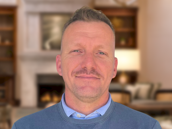 A man with short hair and a mustache wearing a blue sweater and name tag, standing in a well-lit lobby or lounge.