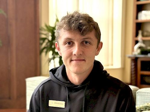 A young man with short hair sits indoors wearing a black hoodie and a name badge, in a warmly lit room with wooden furniture.