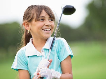 A cheerful young girl in a light blue polo smiles while holding a golf club on a sunny golf course, ready to swing.