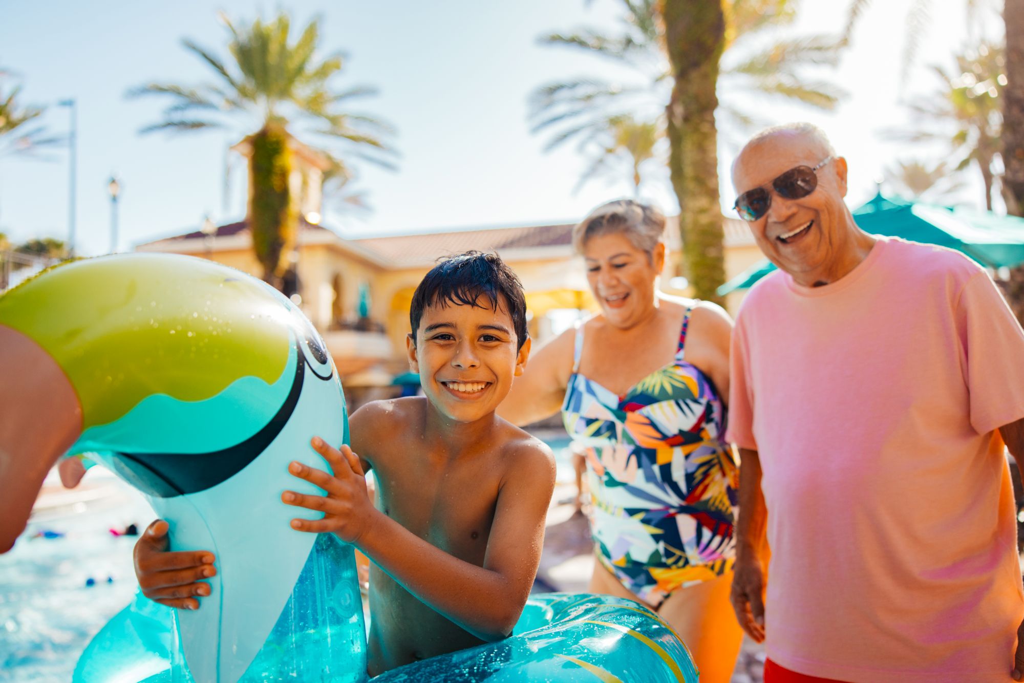 Kids and adults on a sunny pool deck; a boy paddling a blue inflatable dolphin with big smiles, while a smiling couple watches near palm trees.