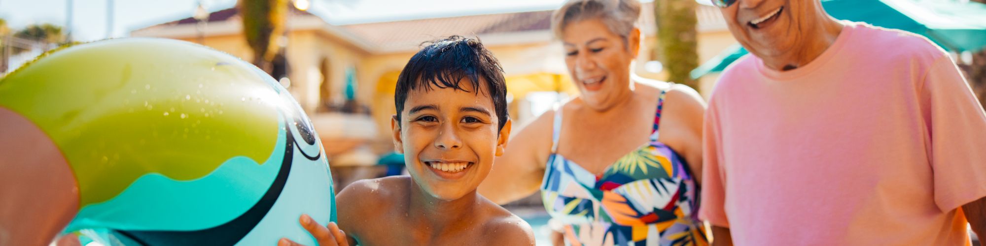 Kids and adults on a sunny pool deck; a boy paddling a blue inflatable dolphin with big smiles, while a smiling couple watches near palm trees.